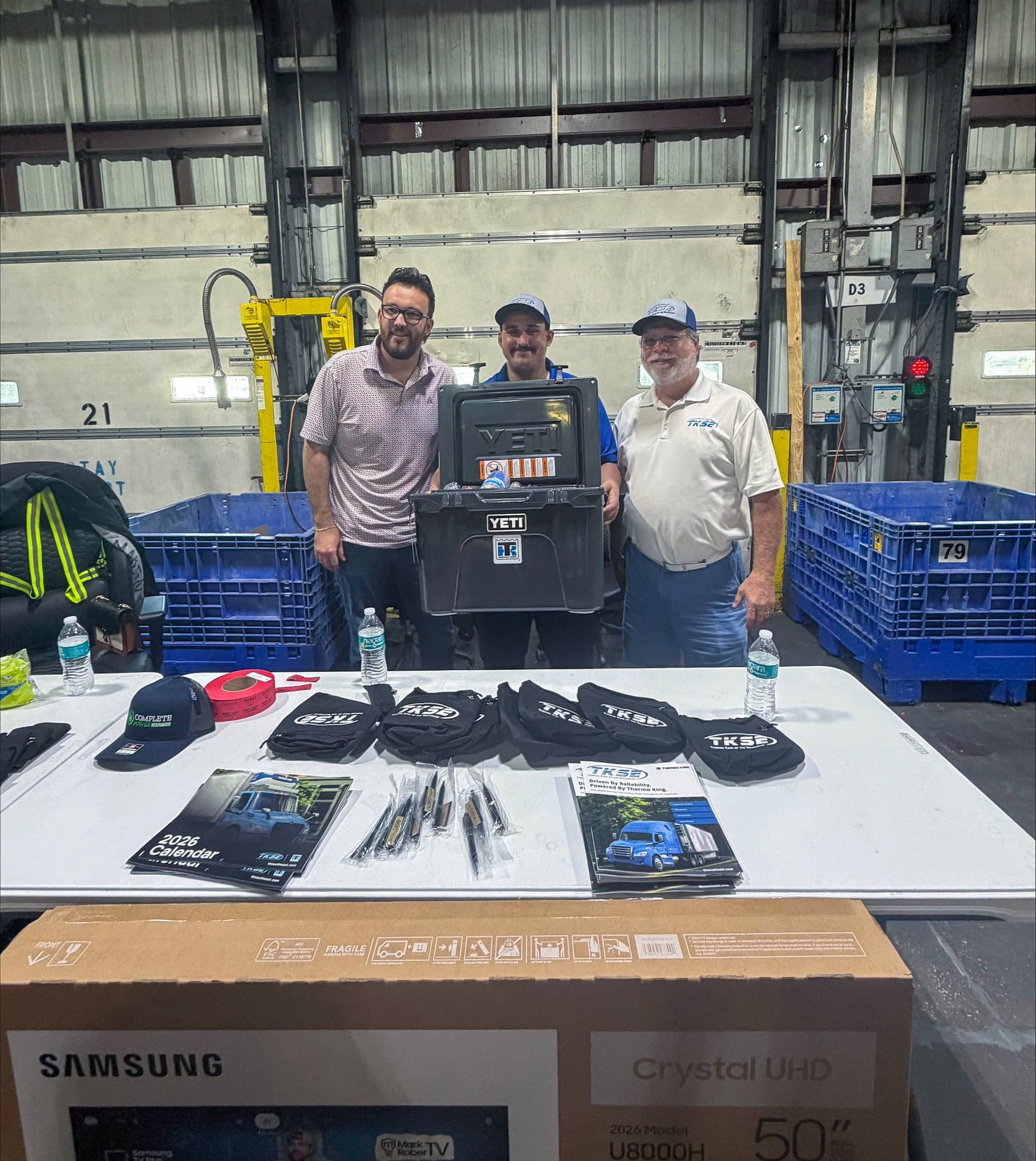 Three people behind a table of TKSE giveaways, with a YETI cooler, TKSE drawstring bags and 2026 calendars, in a warehouse