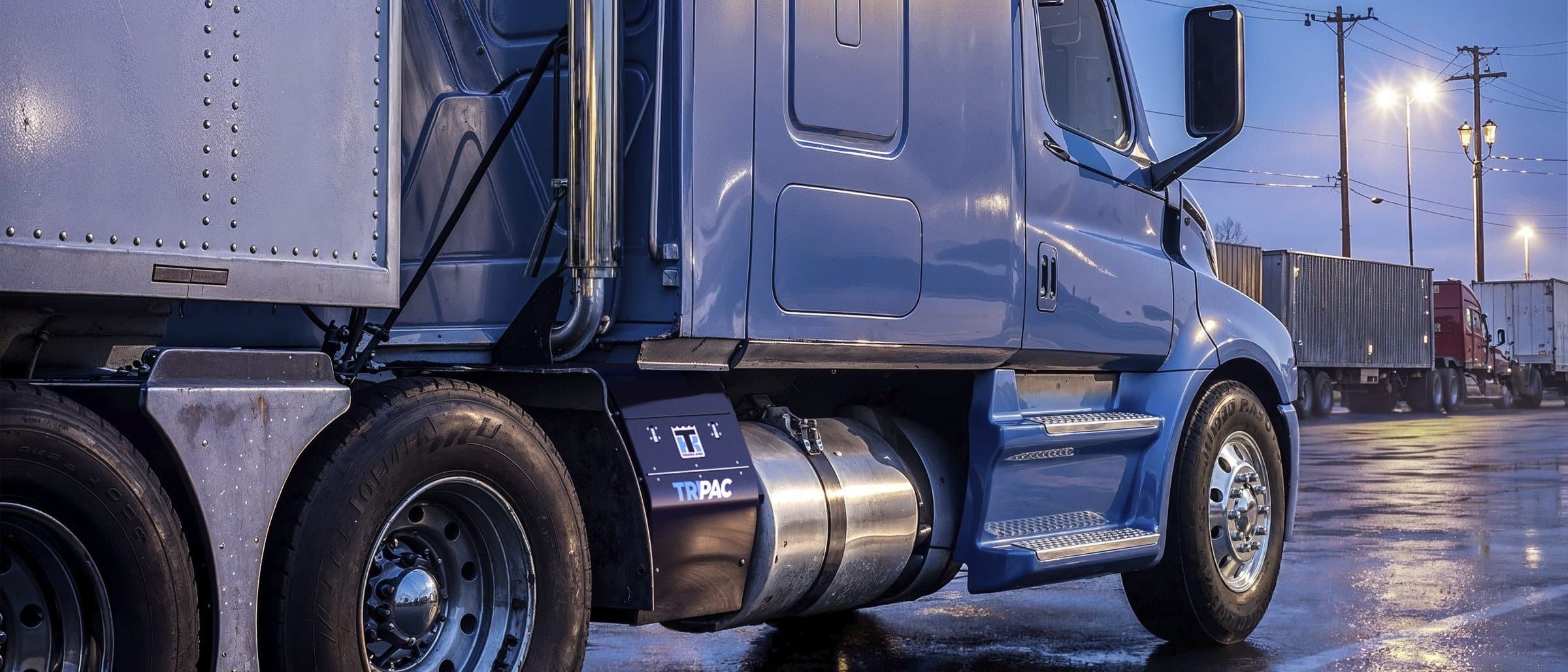 A blue tractor parked at a truck stop at dusk, with a TriPac APU mounted behind its fuel tank
