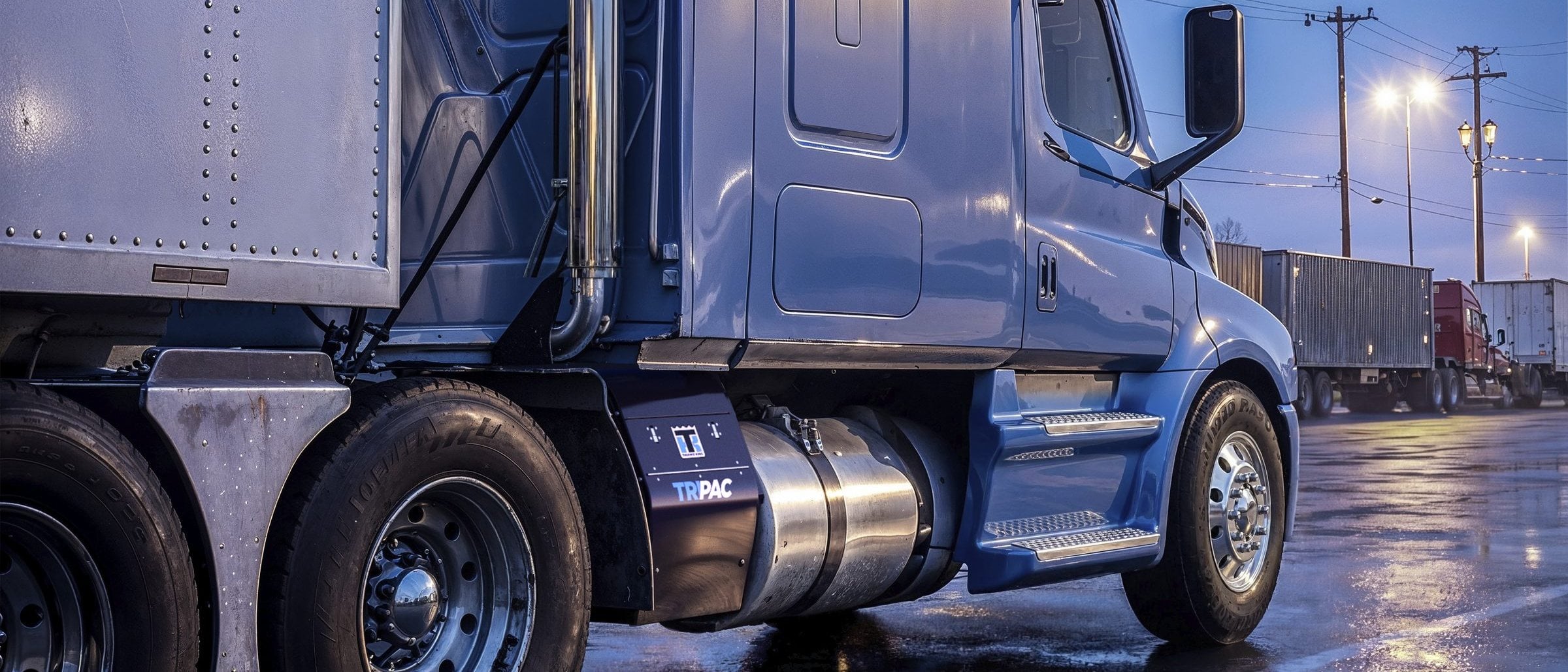 A blue tractor parked at a truck stop at dusk, with a TriPac APU mounted behind its fuel tank