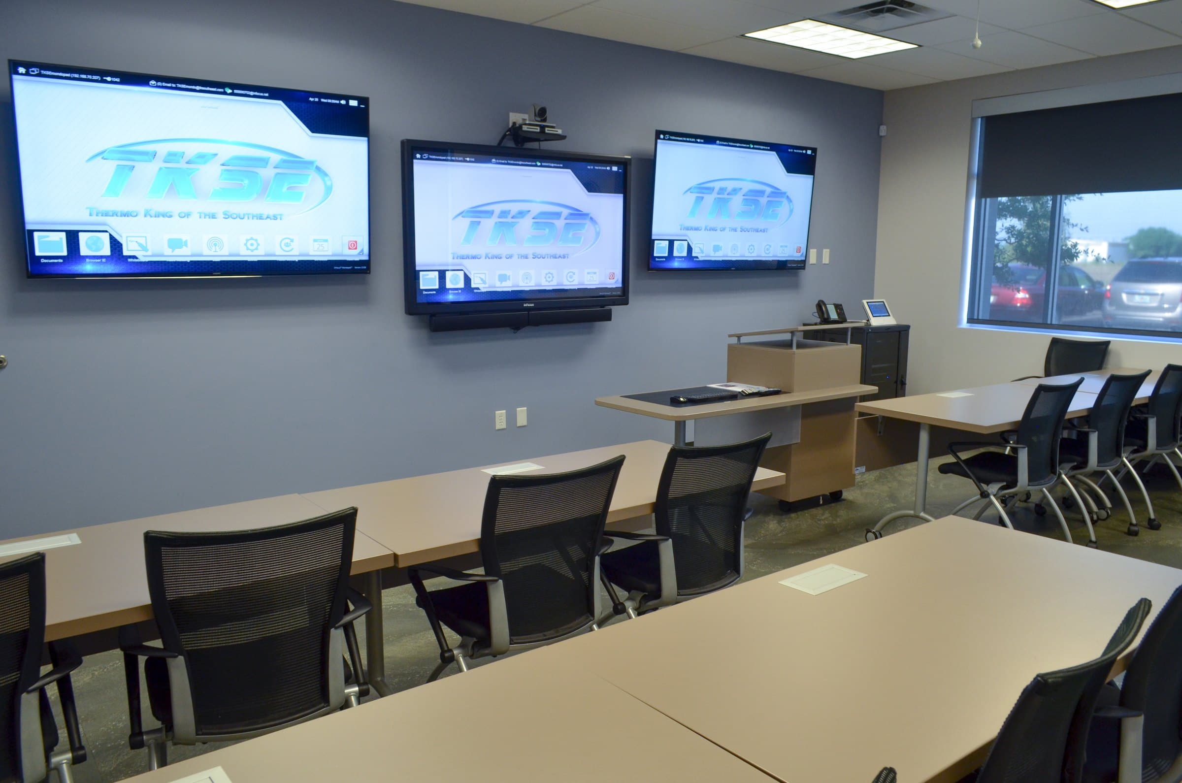 TKSE's training center: three large screens on the wall with a camera above the middle one, and rows of desks with mesh chairs